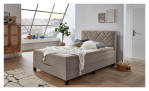 Boxspringbett Josh aus einem Mikrofaser Bezug in Beige mit schwarzen Füßen. Auf einem dekorierten Hintergrund.